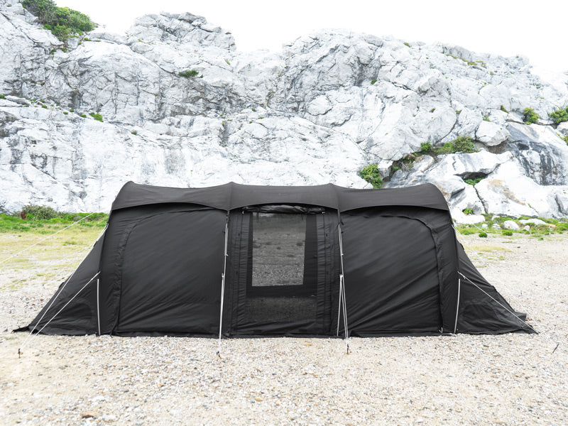 A large black camping tent pitched on a gravel ground with a rocky cliff face in the background.