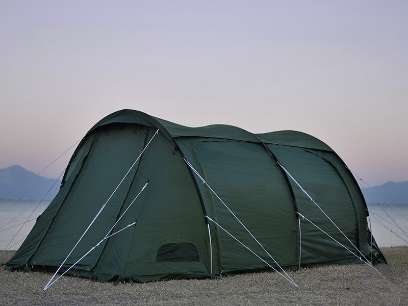 A dark green dome-style camping tent pitched on a sandy shore with water and distant mountains under a pale twilight sky.
