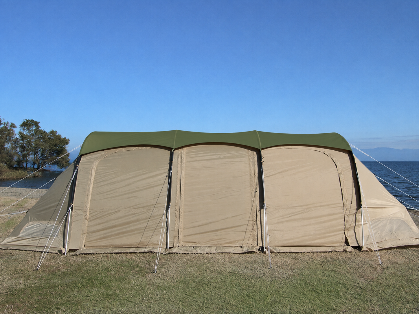 A large beige and olive green camping tent pitched on grass near a body of water under a clear blue sky.