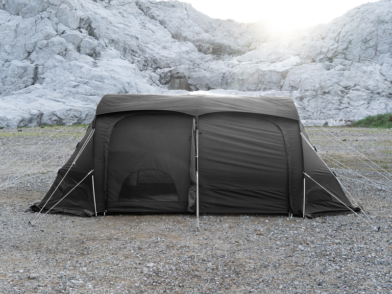 A large black camping tent pitched on a gravel ground with a rocky mountain backdrop under bright sunlight.