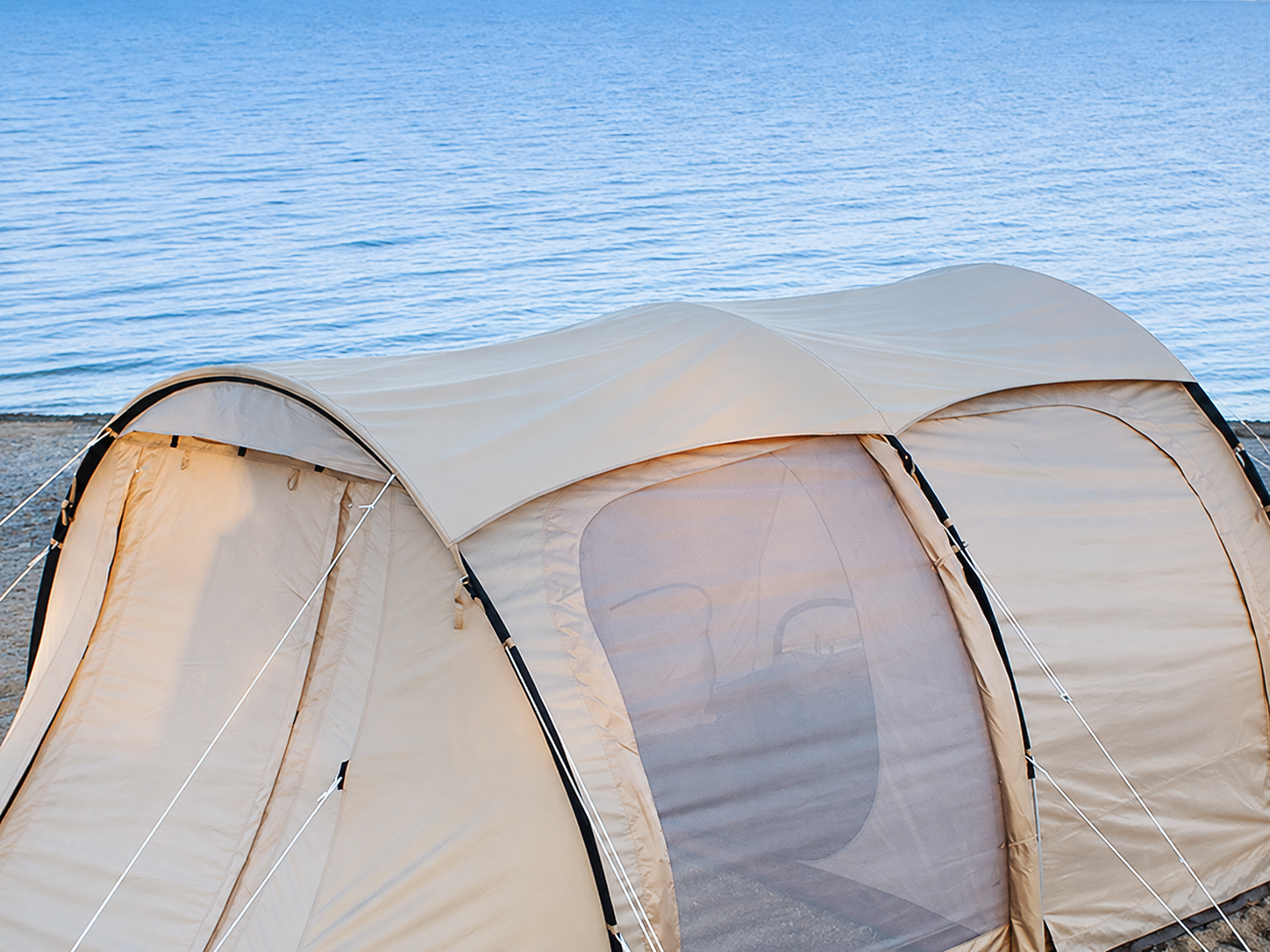 A beige dome camping tent pitched on a beach with blue water in the background.