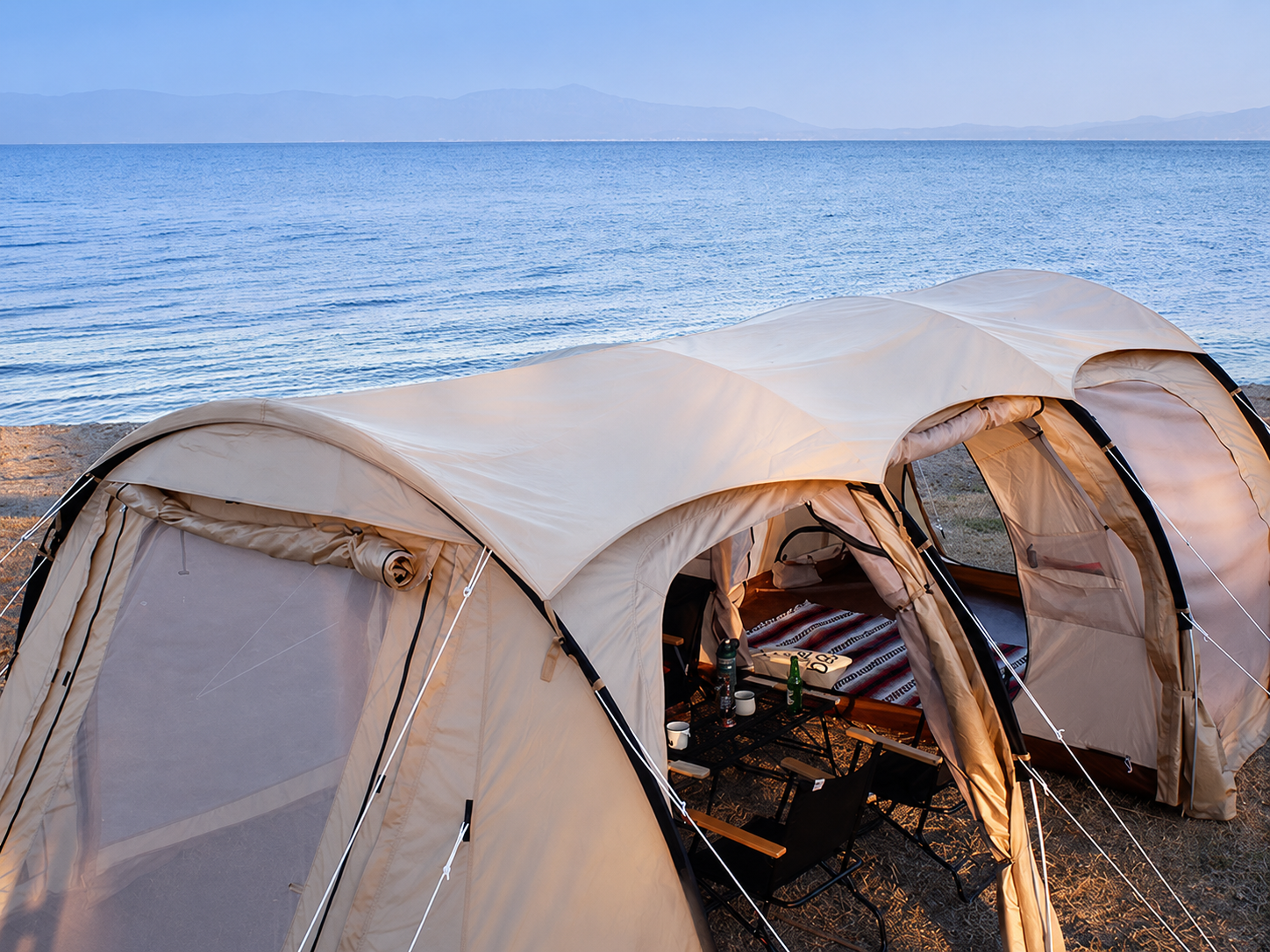 A large beige dome camping tent pitched on a lakeshore with the blue water and distant mountains in the background. The tent is open, showing a sleeping bag, pillows, and a small table inside.