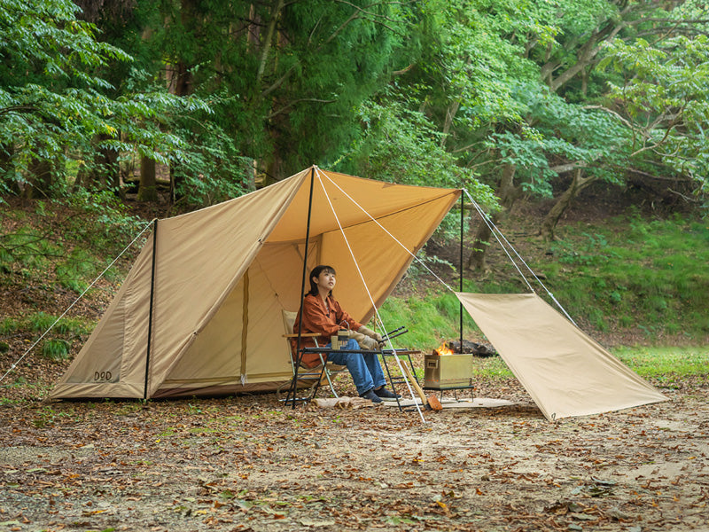 A tan camping tarp shelter set up in a forest clearing, with a person sitting inside it at a small table and a fire pit. A matching beige ground sheet is laid out on the ground beside it.