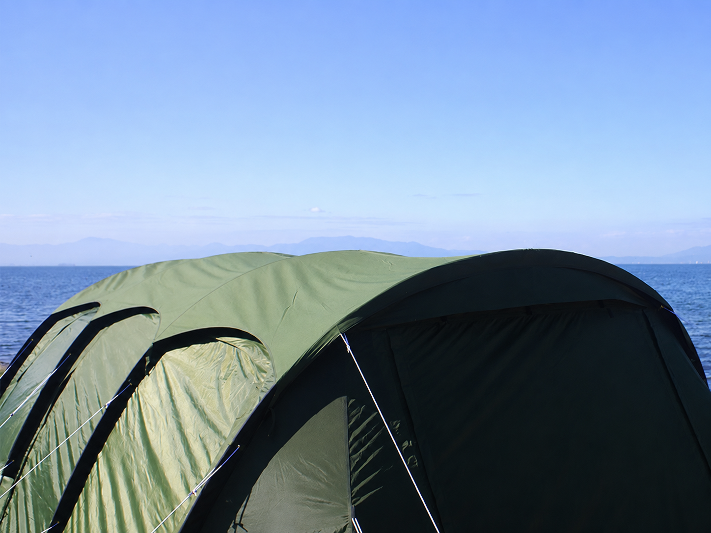 An olive green dome tent pitched outdoors with the ocean and blue sky in the background.