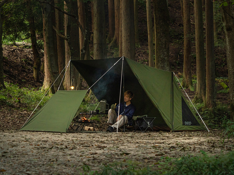 An olive green camping tarp tent with a black fly sheet is pitched in a forest clearing, with a person sitting inside near a small campfire. The tent has a green front panel, a black fly sheet, and white guy lines, with the brand name 'DODO' visible on the lower right corner.