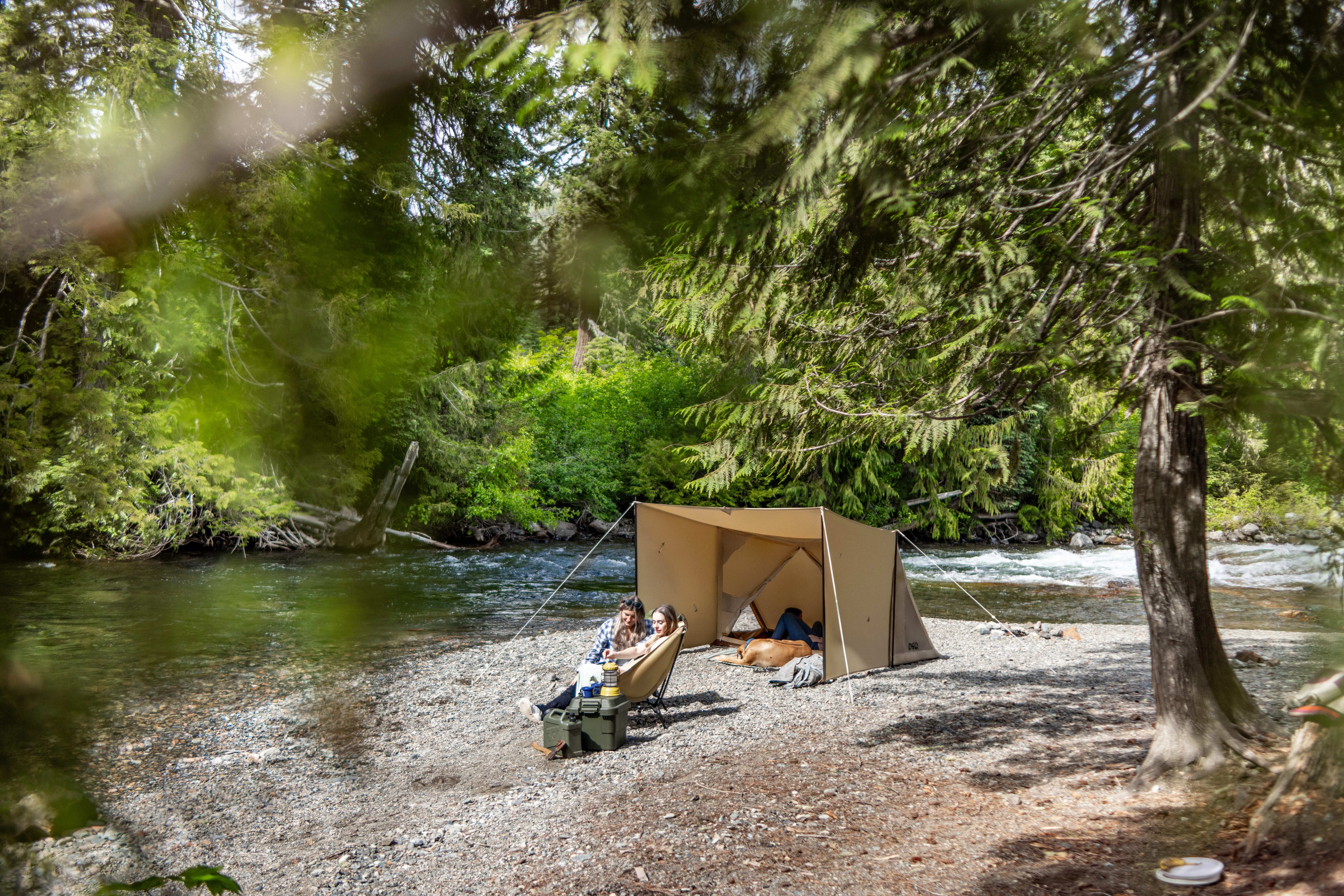 A tan camping tarp shelter set up on a rocky riverbank in a forest, with two people sitting in lounge chairs nearby and a river flowing behind it.