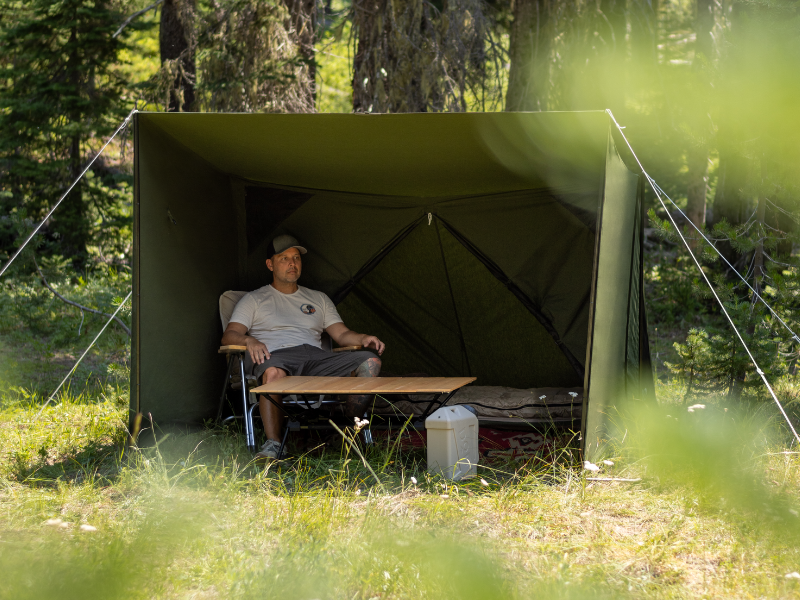 A man sits inside an olive green camping shelter tent set up in a grassy forest clearing, with trees and blurred foliage in the background.