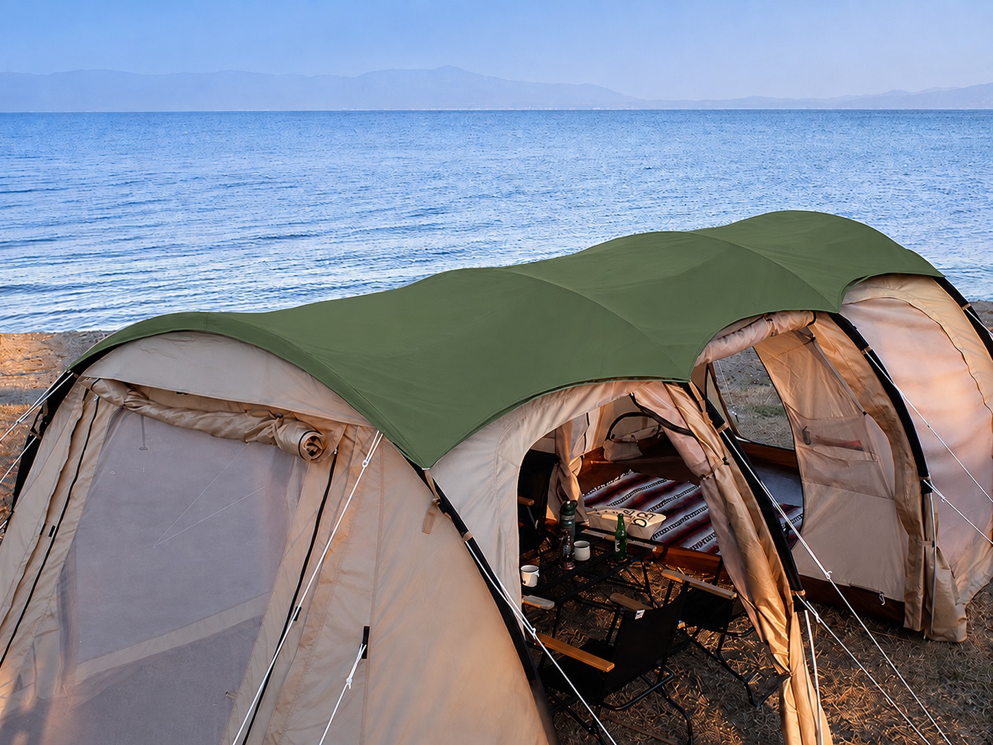 A large dome-style camping tent pitched on a beach or shoreline with the sea and distant mountains in the background. The tent has a green roof and beige/tan walls and mesh doors, with a bed and small table visible inside.